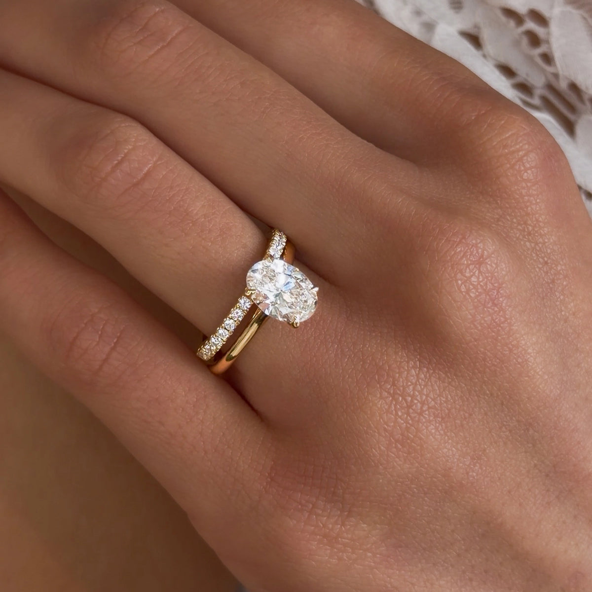  Close-up van een gouden verlovingsring van Aurore Jewelry met een ovale diamant en kleine diamanten op de ringband, gedragen om de ringvinger.